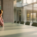 Eine Frau sitzt allein an einem langen, leeren Konferenztisch in einem modernen Glasbüro, Nachmittagslicht fällt durchs Fenster – Symbolbild für das Gefühl, einen sicheren Job zu haben, aber unzufrieden zu sein. KI-generiertes Bild.